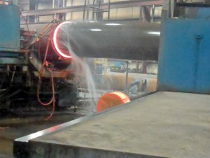 Bending a large pipe at the CBI plant. The glowing band is where the pipe is heated by electric current induction, then bent by the machine at right. Water is sprayed on the already bent portion to prevent over bending.